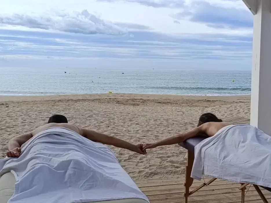 Una pareja frente el mar estirados sobre dos camillas de Masaje Californiano. Se están cogiendo de la mano antes de recibir un masaje en pareja.
