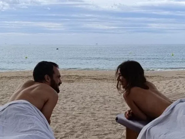 Una pareja frente el mar estirados sobre dos camillas de masaje californiano. Están sonriendo tras recibir un masaje en pareja.